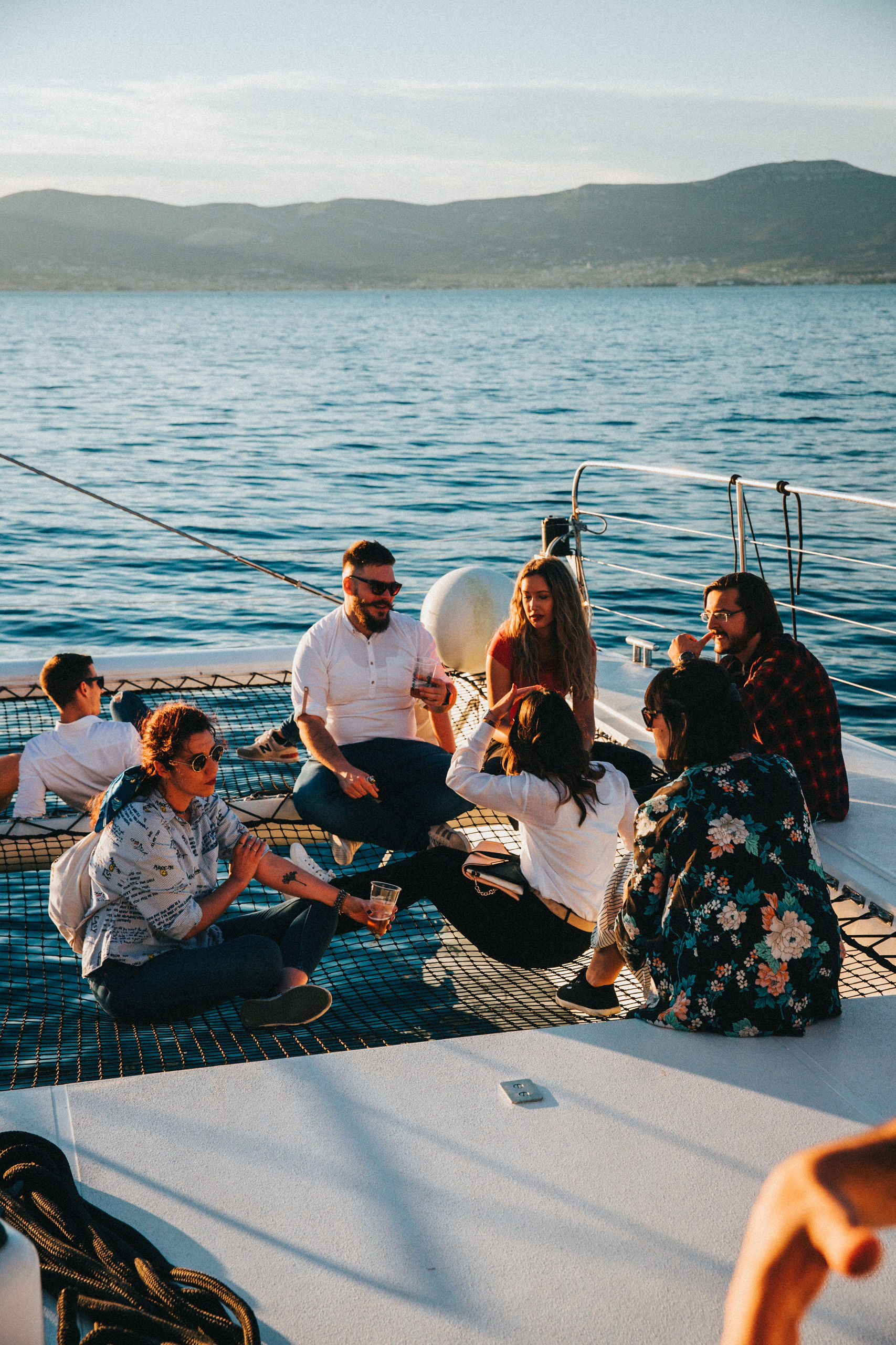 Guests relaxing on catamaran bow net at golden hour with Adriatic mountains behind, yacht party by Cosmic Production