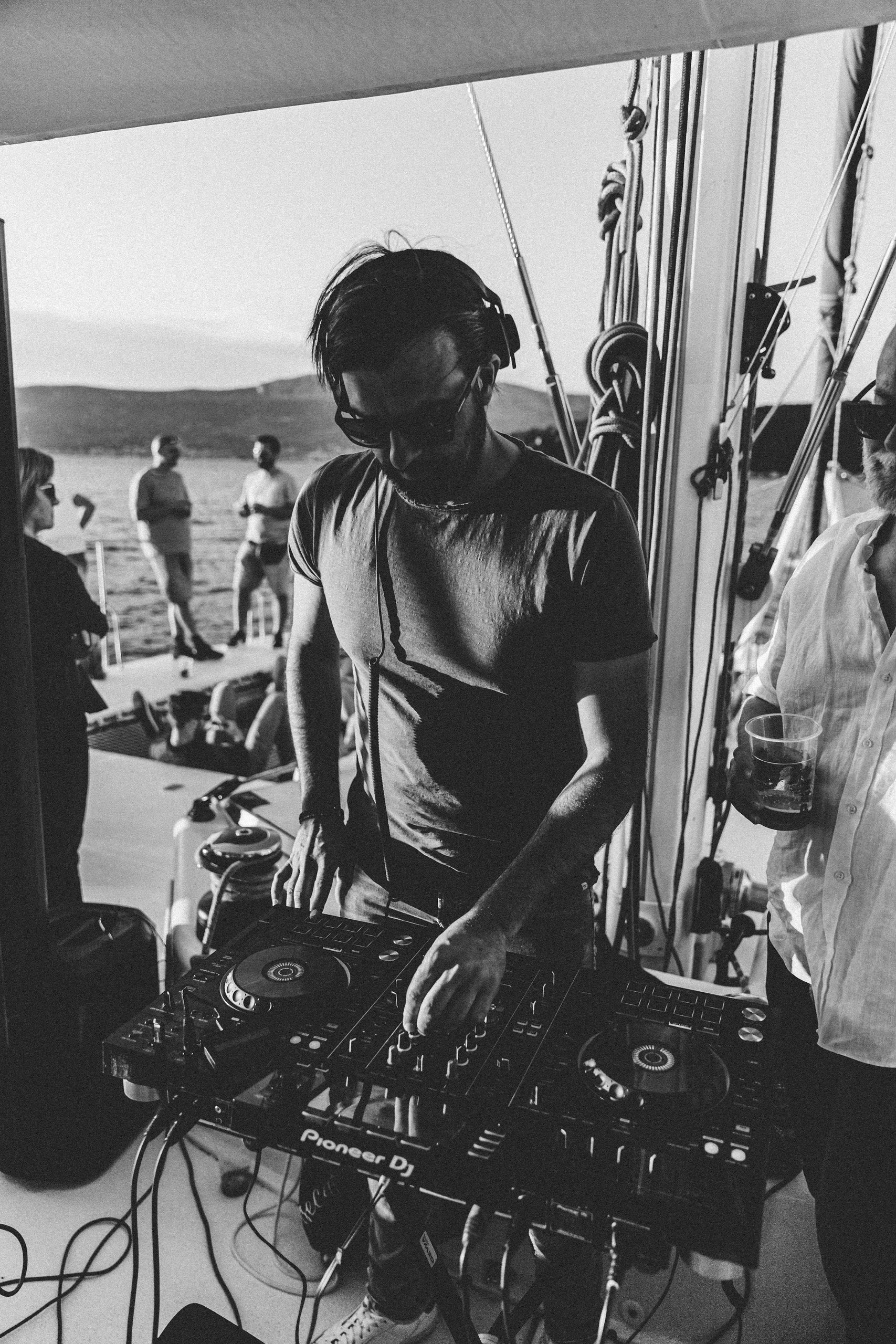 DJ Lomax performing at Pioneer DJ controller on catamaran with rigging and sails framing the shot