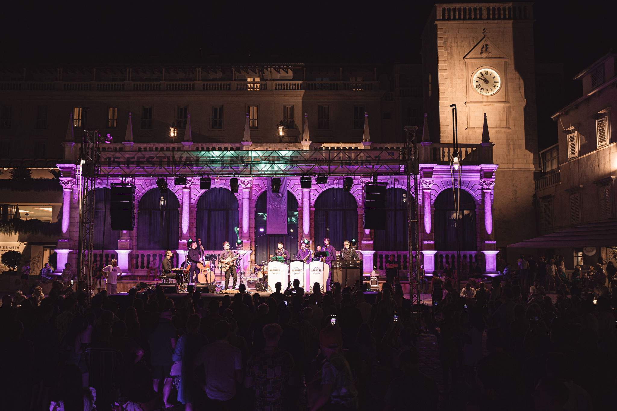 Live band performing on stage with purple-lit loggia arches, speaker stacks, and large crowd at Hvar Town square