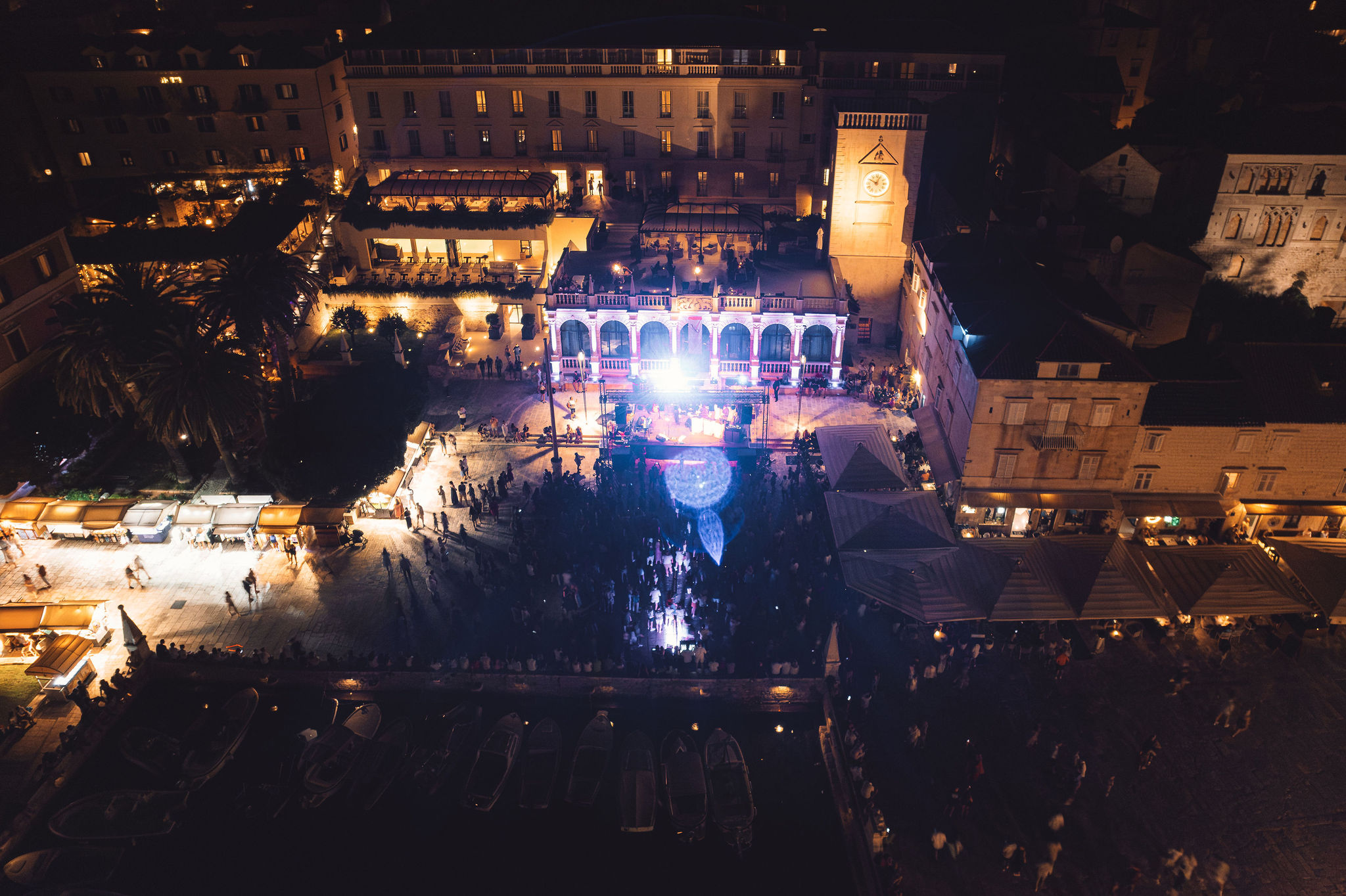 Aerial night view of full concert production on Hvar Town main square with stage, crowd, harbor, and clock tower