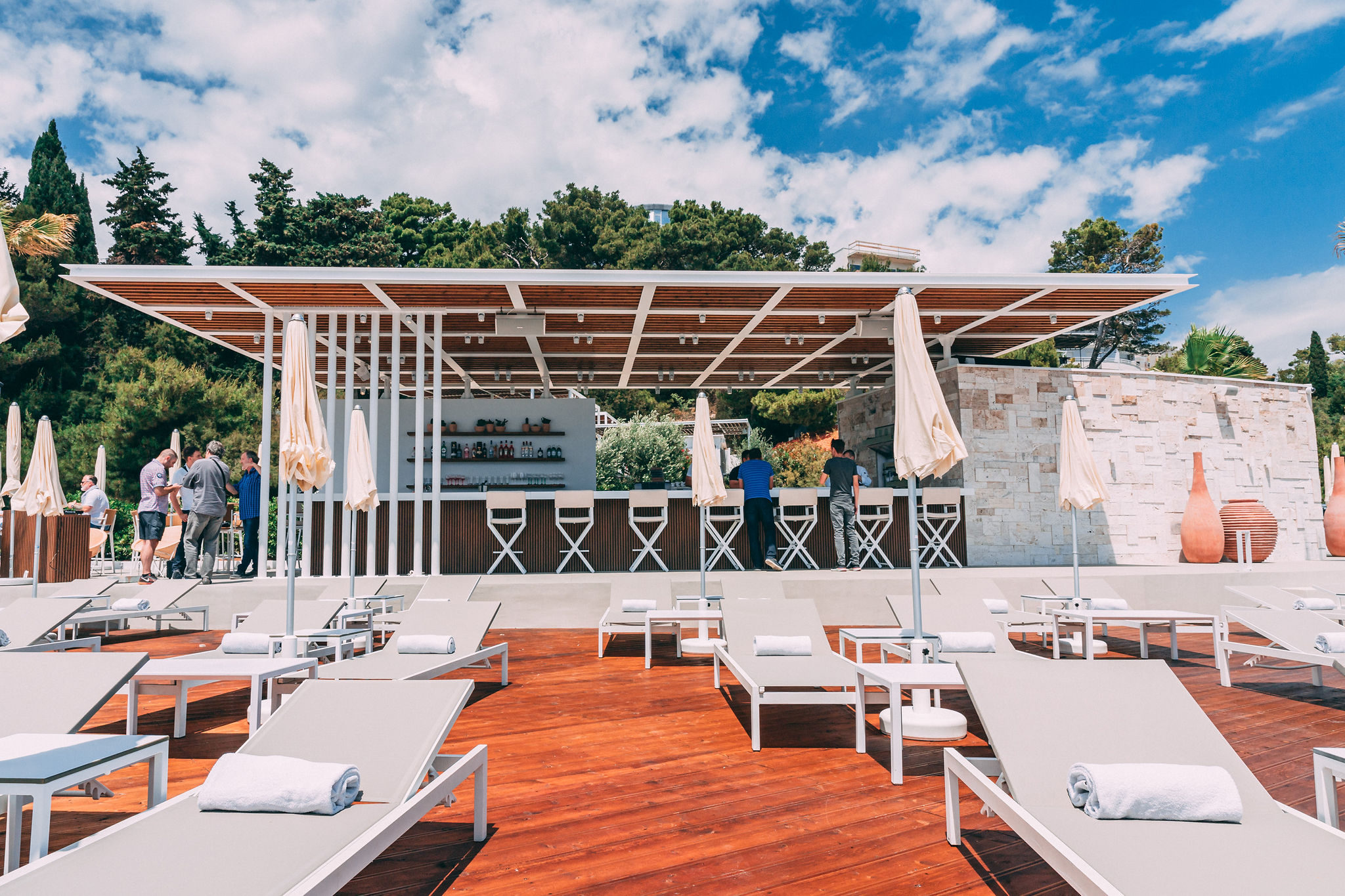 Wide establishing shot of Mistral Beach Club showing DJ booth with Pioneer equipment, ceiling-mounted speakers in pergola grid, bar area, sun loungers, and Adriatic Sea at Radisson Blu Split