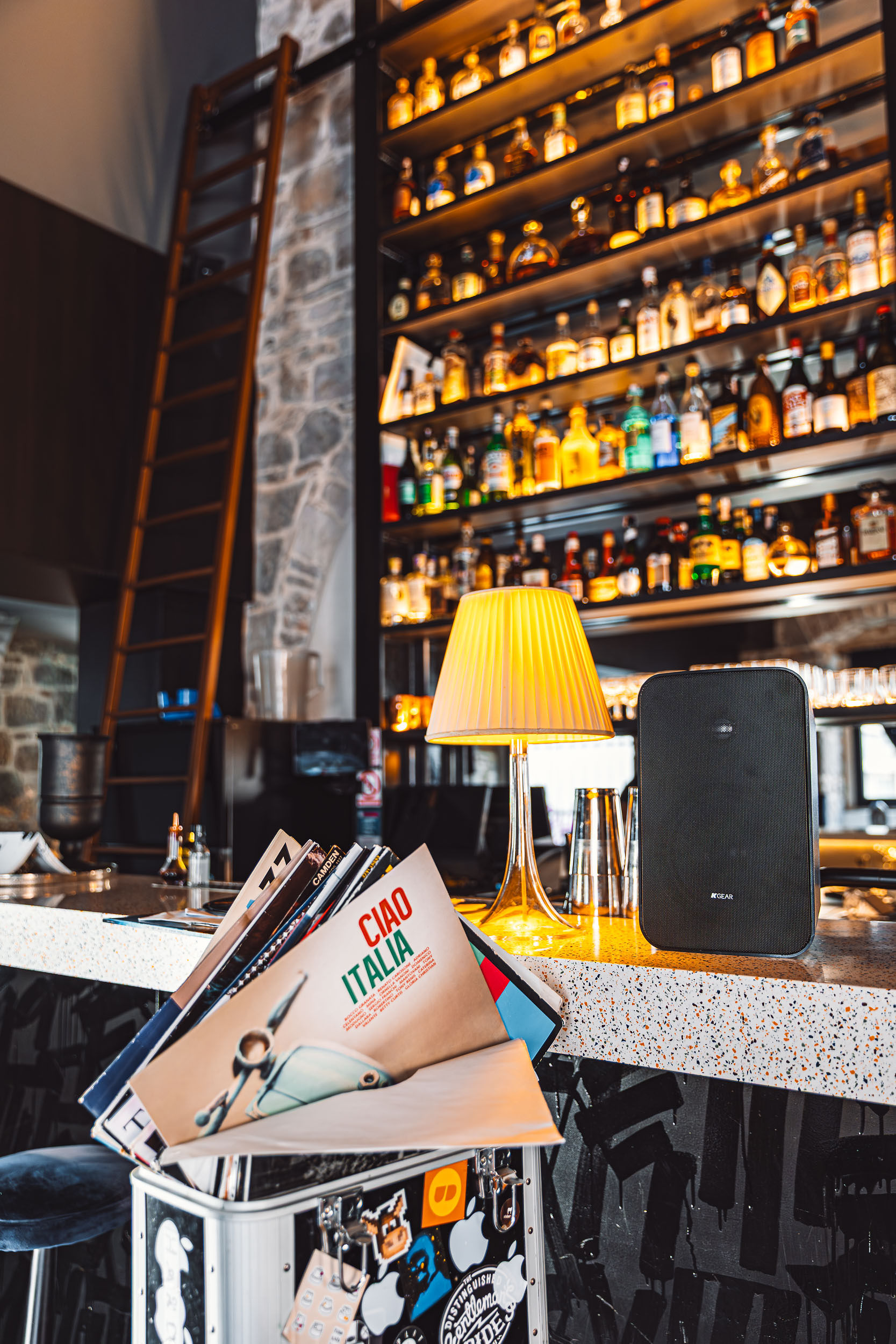 Vinyl records alongside K-Gear speaker with illuminated bottle shelves creating warm mood at Bar Sistema Split